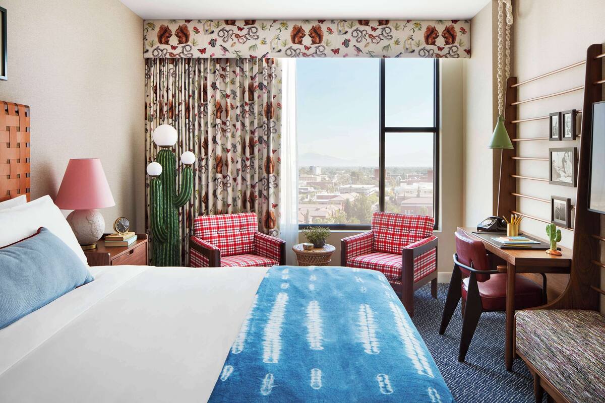 A hotel room with a large bed, two red checkered chairs, a desk with a lamp, a cactus, and a view of a cityscape.
