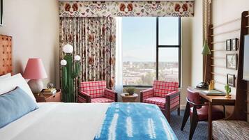 Habitación de hotel con una cama grande, dos sillas rojas de cuadros, un escritorio con lámpara, un cactus y vistas a un paisaje urbano.