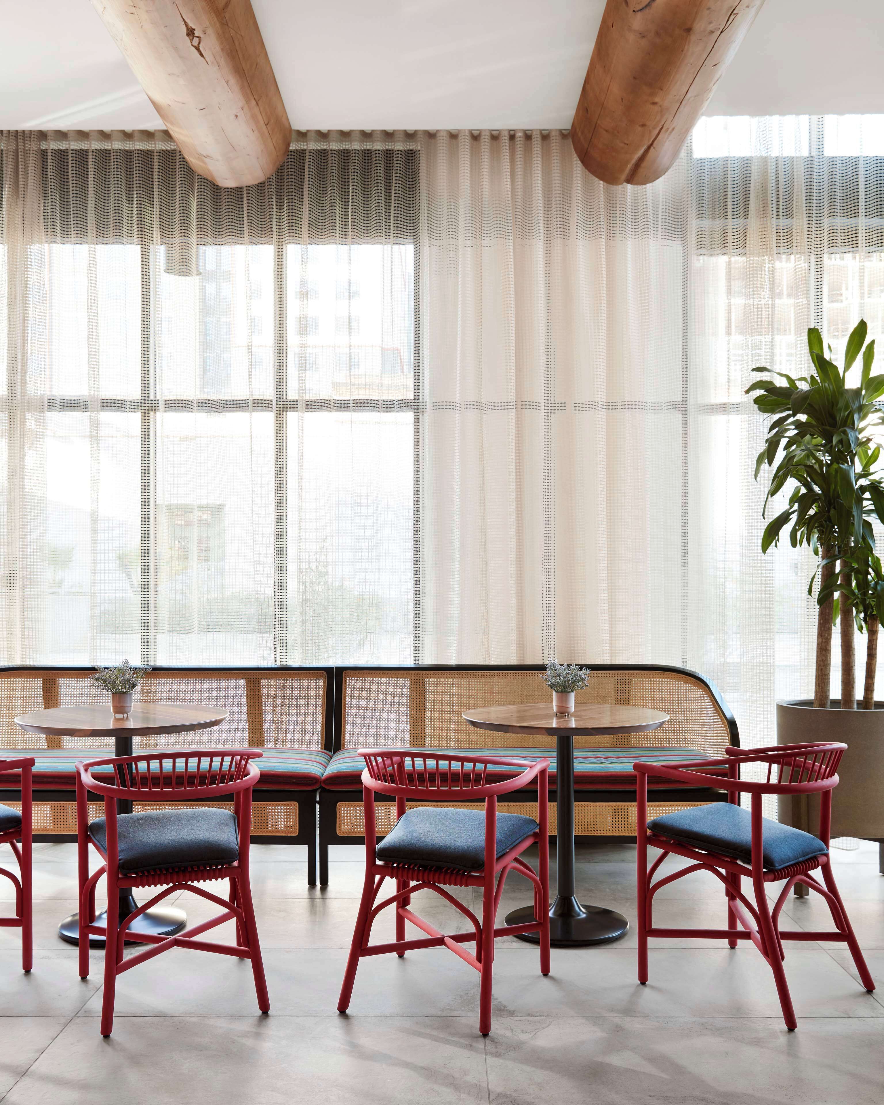 A modern interior with a seating area featuring red chairs and a round table, large windows with sheer curtains, and a potted plant.