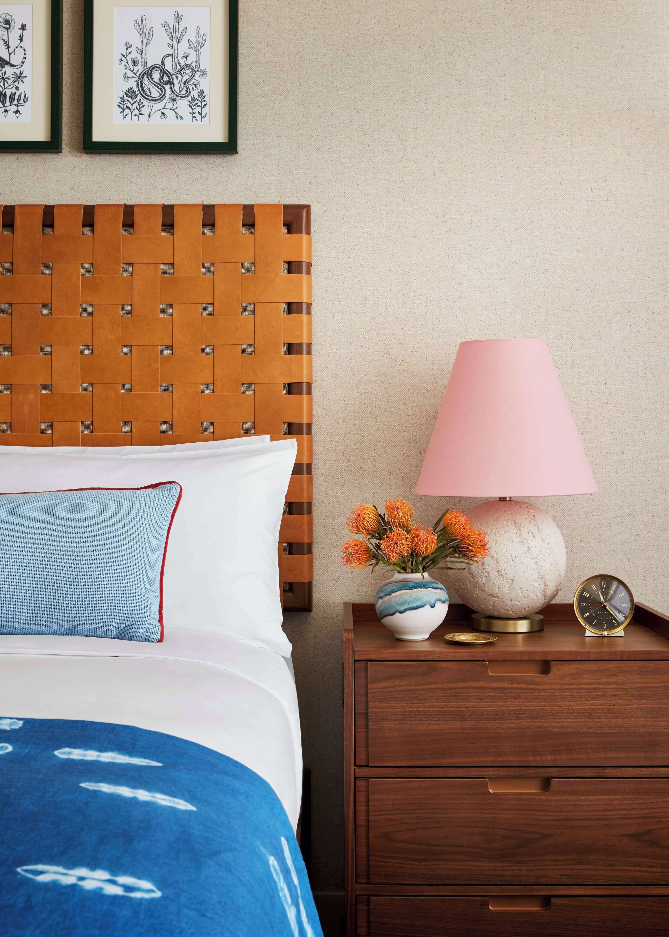 Un lit bien fait, avec une couverture bleue, une tête de lit en bois et une table de chevet sur laquelle se trouvent une lampe rose, un vase de fleurs et une horloge.