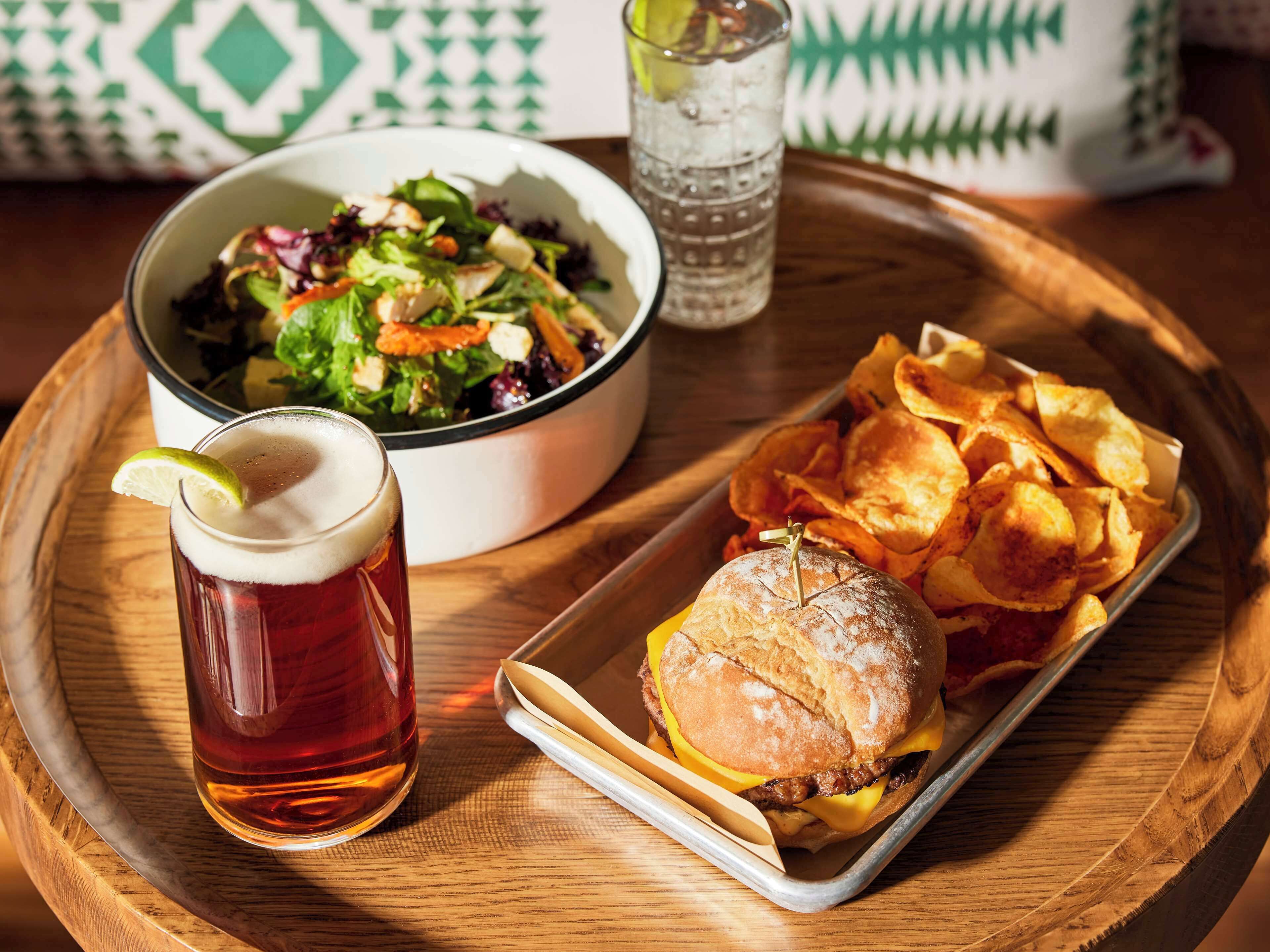 A meal tray with a burger, chips, and a glass of beer, along with a bowl of salad.