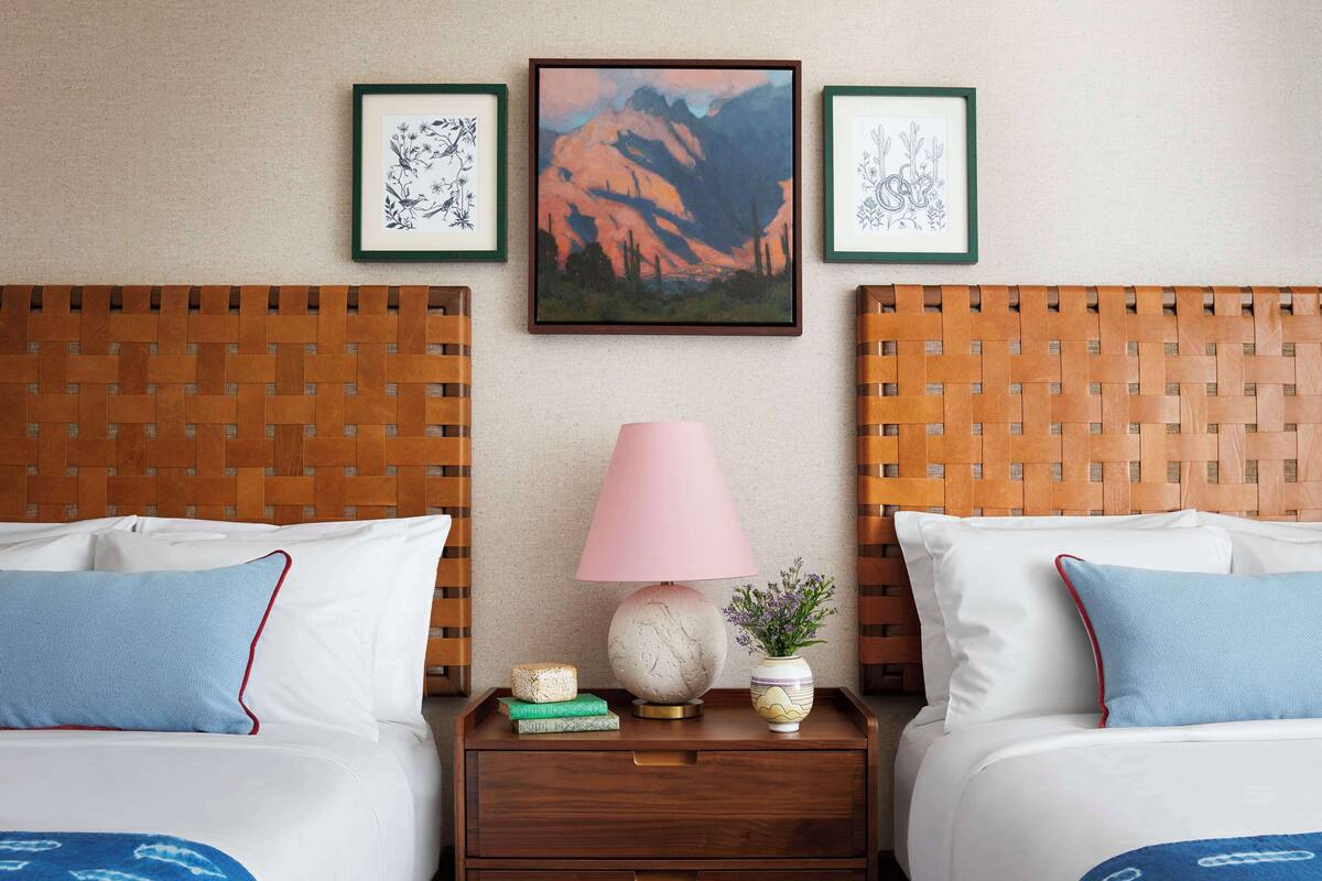 A hotel room with two beds, a nightstand with a lamp, framed artwork on the wall, and a woven headboard.