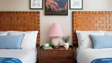 Habitación de hotel con dos camas, una mesita de noche con lámpara, una obra de arte enmarcada en la pared y un cabecero tejido.