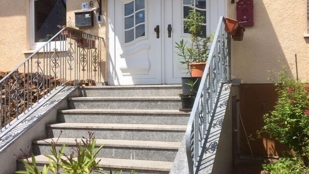 A building with a white double door, a staircase with metal railings, and potted plants around the entrance. - Ferienwohnung Albert, 2 Schlafzimmer, 90 Quadratmeter (Hauenstein)