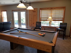 Une table de billard avec les queues et les billes, deux chaises, une lampe et une fenêtre avec des stores.