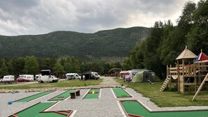 Um camping com várias caravanas de telhado verde, uma estrutura de recreação de madeira e vista para uma cadeia montanhosa arborizada.