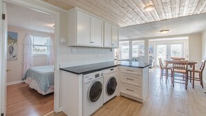 A compact kitchen with a washing machine, a dining area with a table and chairs, and a bedroom with a bed and curtains.