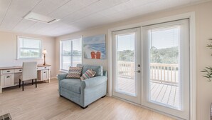 A living room with a light blue sofa, a desk, and a chair. There are two windows with blinds, a painting on the wall, and a door leading to a balcony.