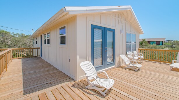 A small, single-story building with a deck, white walls, blue shutters, and a few white chairs.