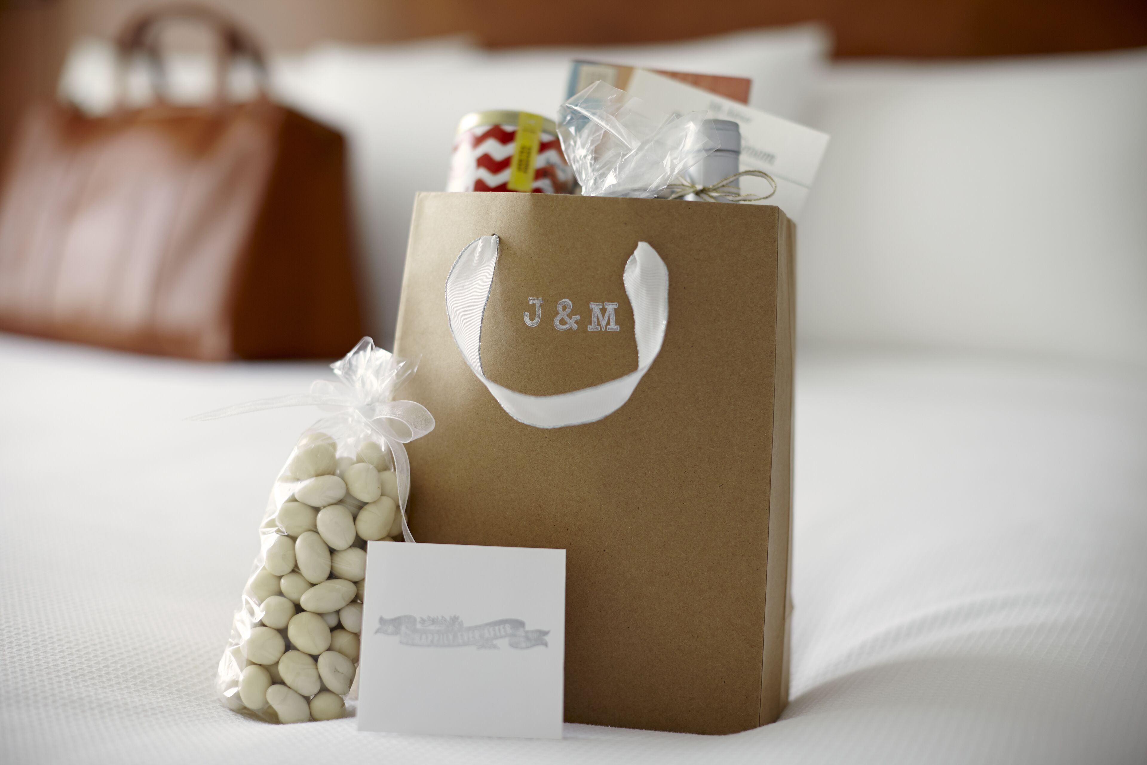 Una bolsa de regalo con la marca J&M, un lazo blanco, una tarjeta pequeña blanca y un paquete de judías blancas sobre una cama con ropa blanca y un bolso de cuero marrón al fondo.