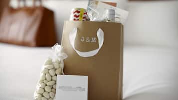 Una bolsa de regalo con la marca J&M, un lazo blanco, una tarjeta pequeña blanca y un paquete de judías blancas sobre una cama con ropa blanca y un bolso de cuero marrón al fondo.