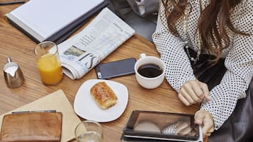 A wooden table with a newspaper, a glass of orange juice, a cup of coffee, a pastry, a wallet, a glass of water, and a tablet.