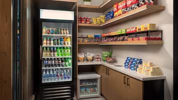 Máquina expendedora llena de diversas bebidas y aperitivos, estantes con productos envasados y un refrigerador pequeño.