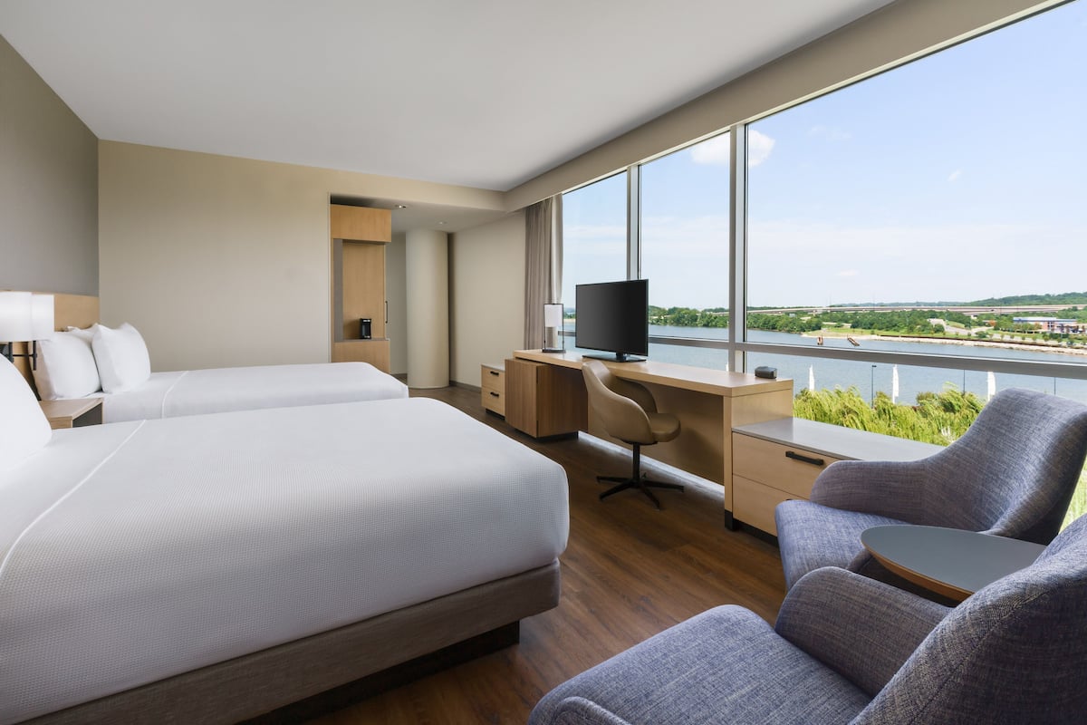 A hotel room with two beds, a desk, a chair, a TV, and a large window with a view of a river and greenery.