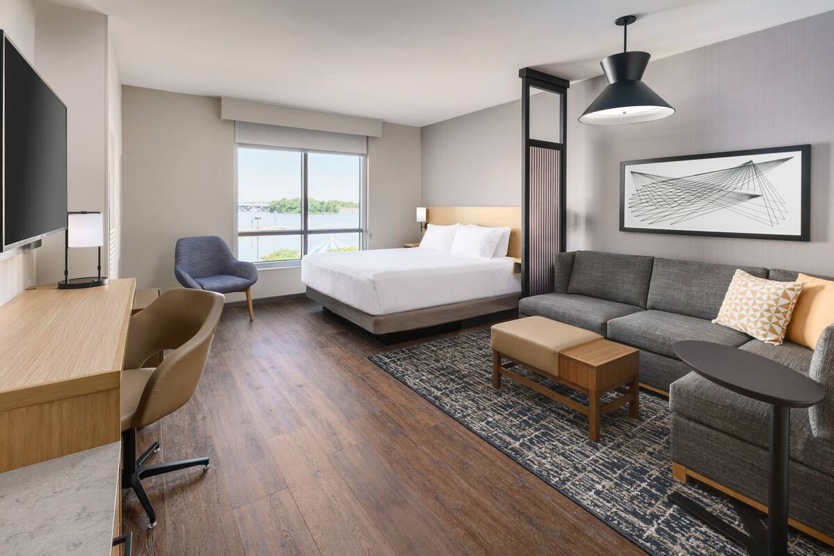 A modern hotel room with a bed, sofa, desk, chair, and a large window offering a view.