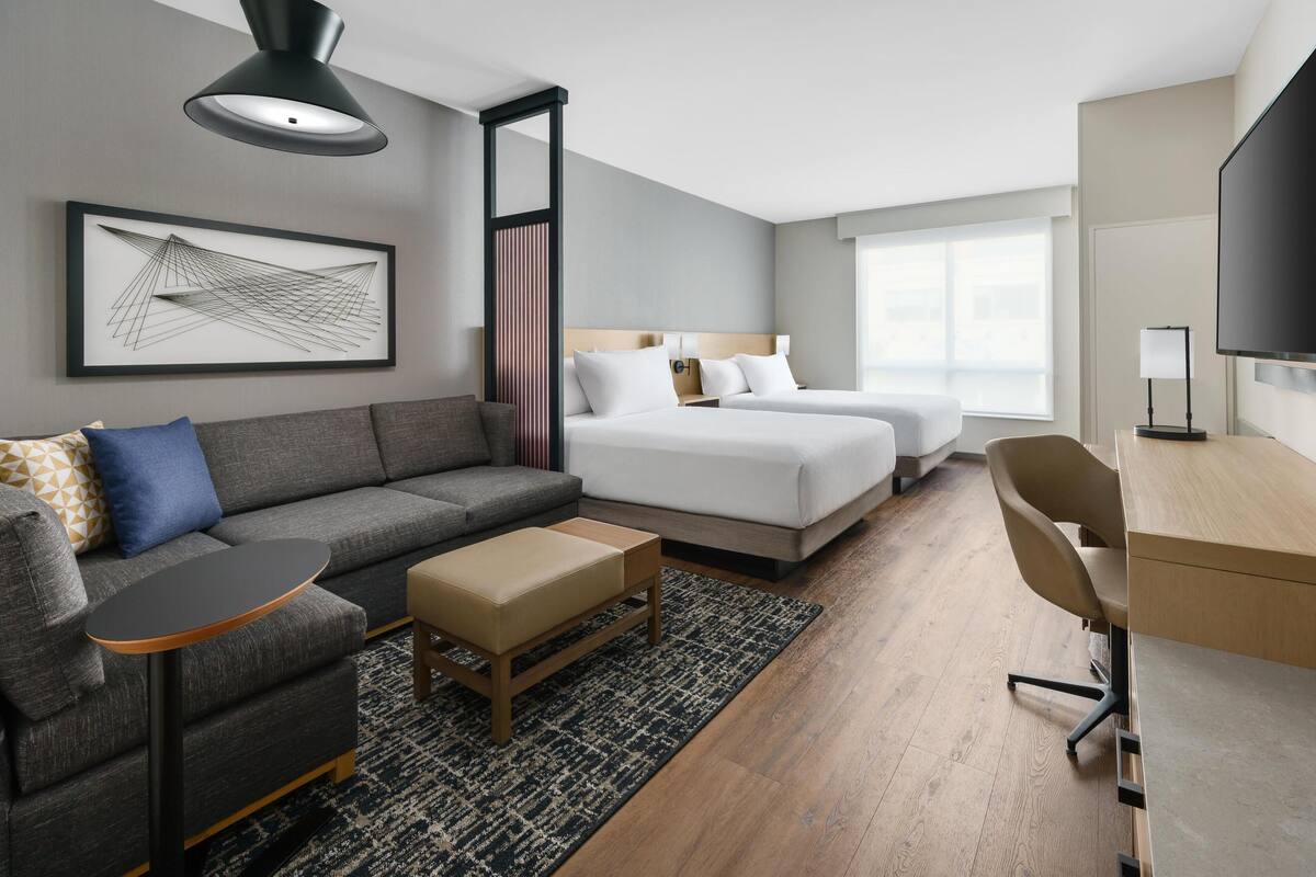 A modern hotel room with a sofa, two beds, a desk, and a TV.