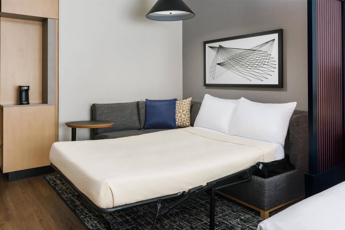 A modern hotel room with a bed, a small table, a sofa, and a coffee machine.