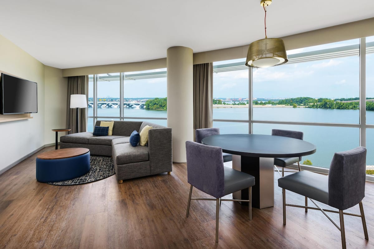 A modern hotel room with a large window, a sofa, a round dining table, and a flat-screen TV.