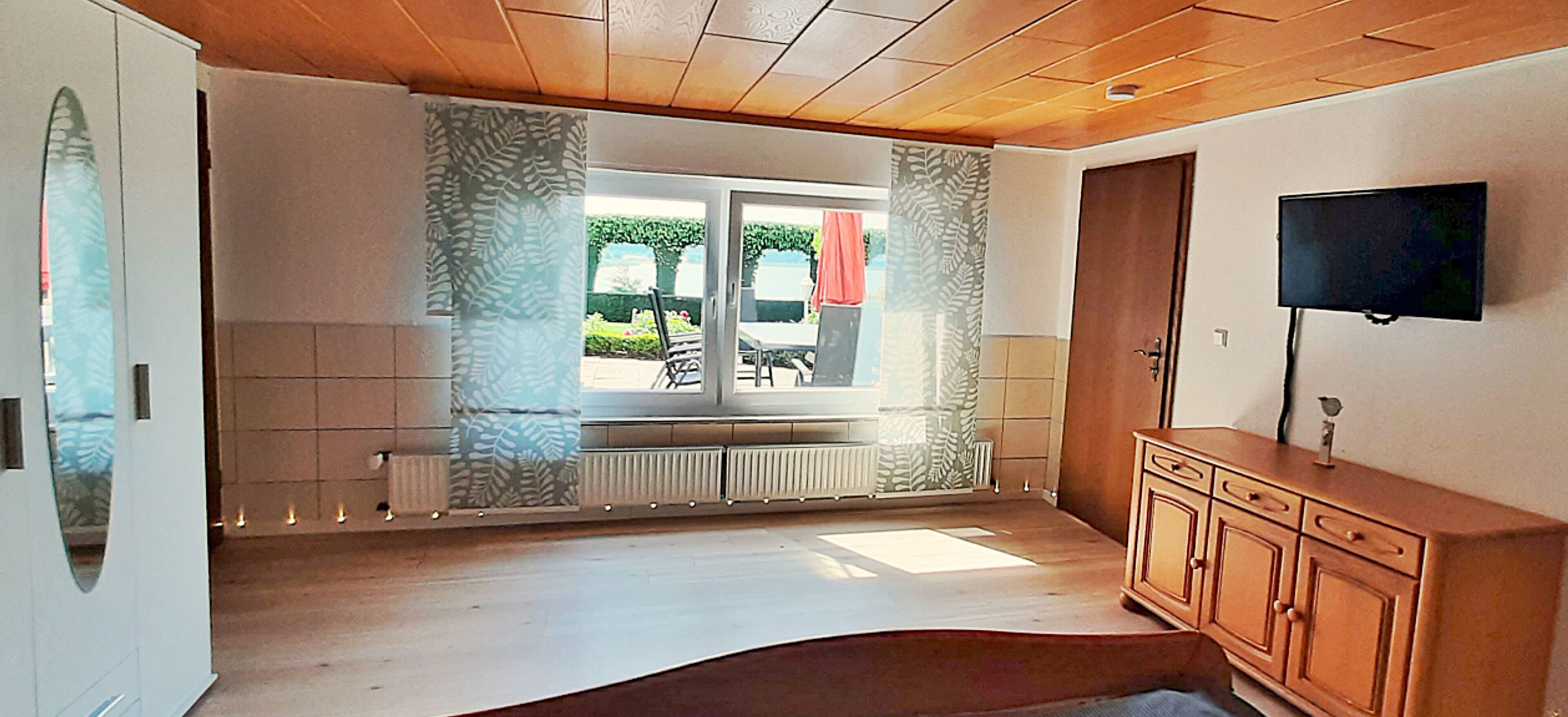 Una habitación con techo de madera, un ventanal con cortinas, un radiador, un televisor montado en la pared y un armario de madera.