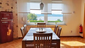 Zona de dinar amb taula, cadires, nevera i finestra amb vista a vegetació i aigua.