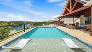 Ein Schwimmbecken mit freiem Blick auf die umliegende Landschaft, eine überdachte Terrasse mit Sitzgelegenheiten und ein Steinhaus mit geneigtem Dach.