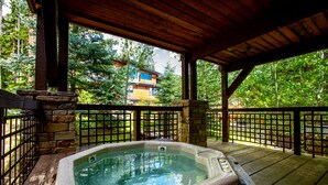 Un jacuzzi en un área exterior cubierta con paredes de piedra y vigas de madera, rodeado por una cerca de metal y abundante vegetación.