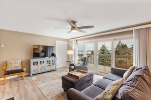 A spacious living room with a sofa, a television, a cabinet, and a balcony with a view of trees.