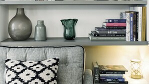Een netjes georganiseerde boekenkast met boeken en decoratieve objecten, een grijze bank met een gestreept kussen, en een kleine tafel met een lamp.