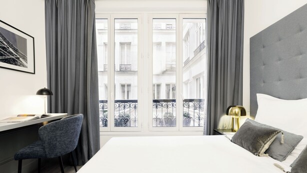 Een moderne hotelkamer met een groot bed, een bureau met stoel en uitzicht op de stad door grote ramen.