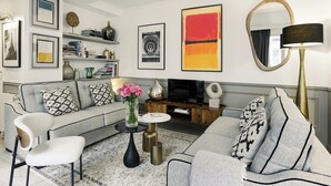 Een moderne woonkamer met een grijze bank, een witte fauteuil, een salontafel en een televisie. Op de muur hangen ingekaderde foto’s, er staat een vaas met bloemen en een lamp.