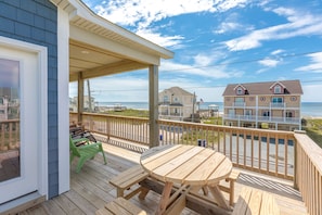 A wooden deck with a table and chairs, overlooking a beach with houses and the ocean.