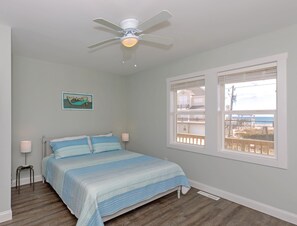 A bedroom with a bed, a ceiling fan, a window with a view of the beach, and a small table with a lamp.