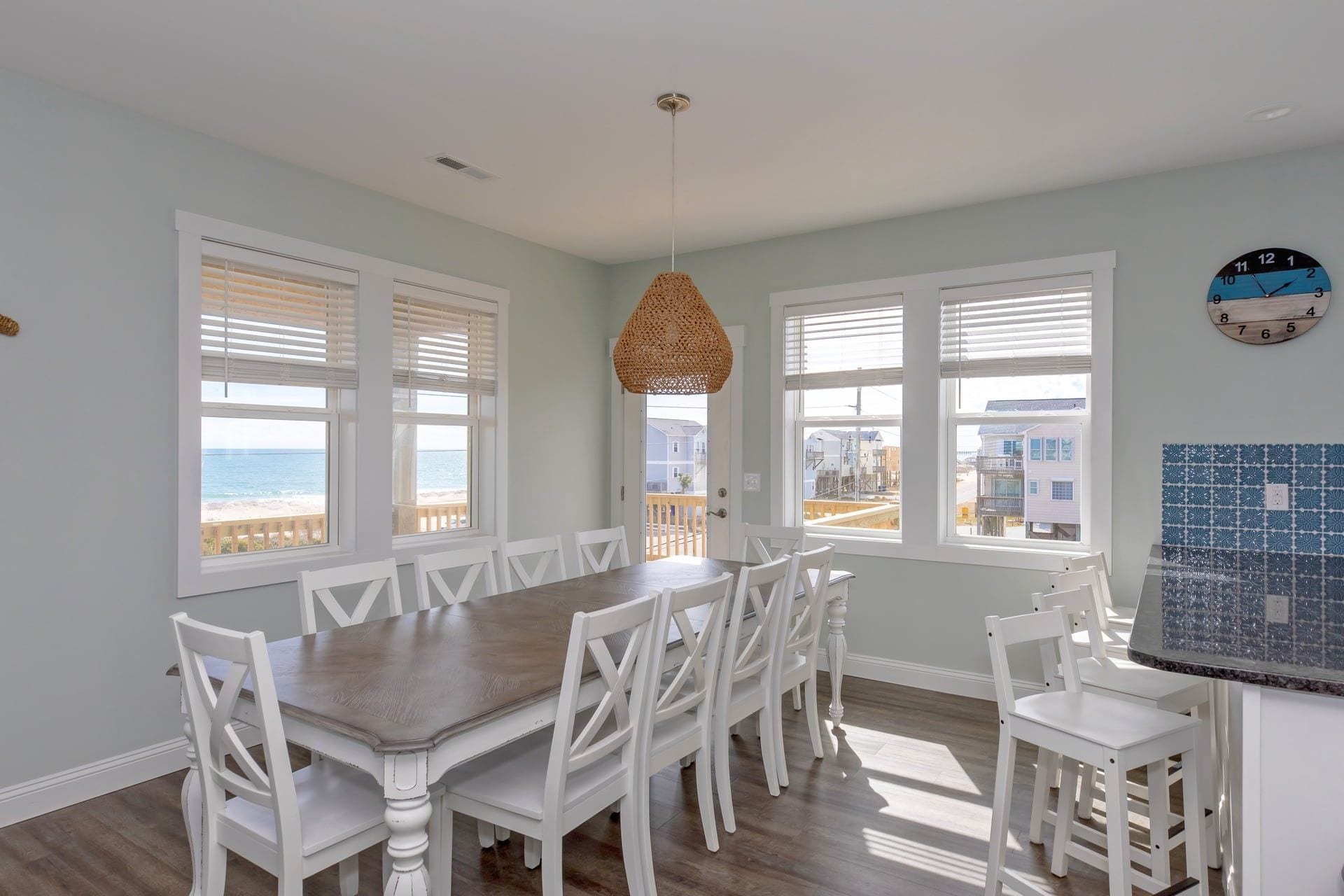 A dining area with a wooden table and white chairs, large windows with blinds, and a view of the ocean.