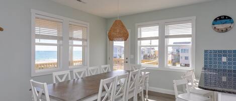 A dining area with a wooden table and white chairs, large windows with blinds, and a view of the ocean.