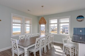 A dining area with a wooden table and white chairs, large windows with blinds, and a view of the ocean.