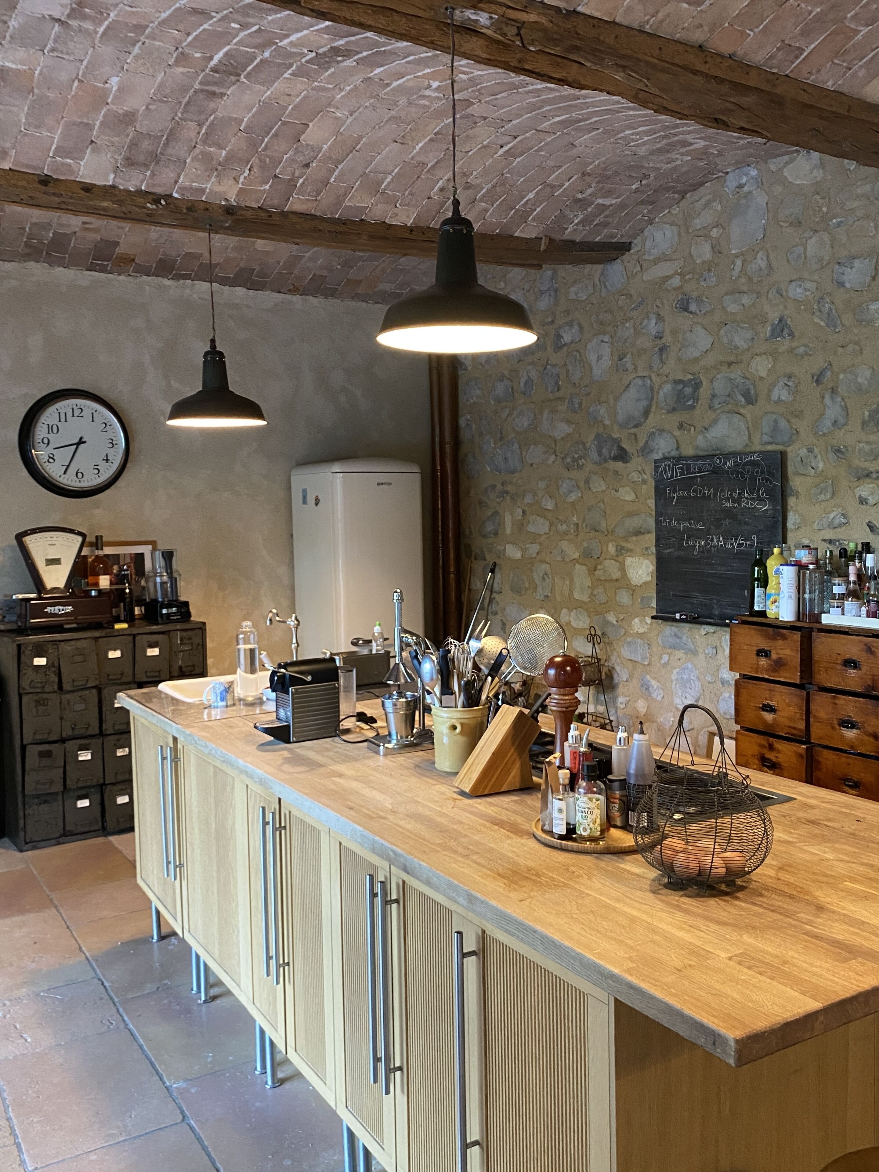 A kitchen with a wooden counter, hanging pendant lights, a clock, a chalkboard with writing, and various kitchen utensils.
