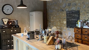 Une cuisine avec un comptoir en bois, des luminaires suspendus, une horloge, un tableau noir avec des inscriptions et divers ustensiles de cuisine.