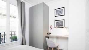 Ein Schlafzimmer mit einem Bett, einem grauen Kleiderschrank, einem Schreibtisch mit Stuhl, einem Blumentopf und zwei gerahmten Bildern an der Wand.