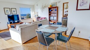 A modern living room with a sofa, dining table, and chairs. There is a TV, bookshelf, and artwork on the walls.