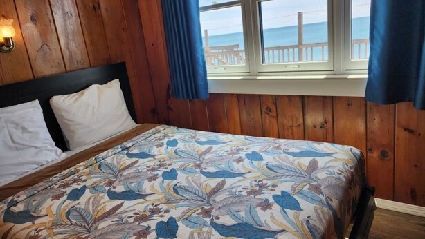 A bedroom with a wooden headboard, a floral bedspread, and a view of the ocean through a window with blue curtains.