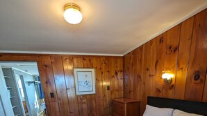A hotel room with wooden panel walls, a bed with floral bedding, two rolled towels, a nightstand, and a framed picture on the wall.