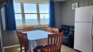 A compact kitchen with a dining table and chairs, a refrigerator, and a view of the sea through large windows.