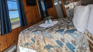 Ein Zimmer mit Holzwänden, einem Bett mit floralem Überwurf, einem Fenster mit Vorhängen, einer Deckenleuchte und einer Küchenzeile.