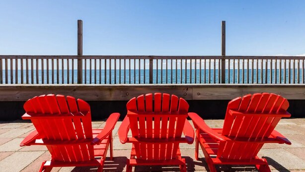 Drei rote Adirondack-Stühle, die auf eine Holzterrasse mit Geländer und Blick auf ein Gewässer zeigen.