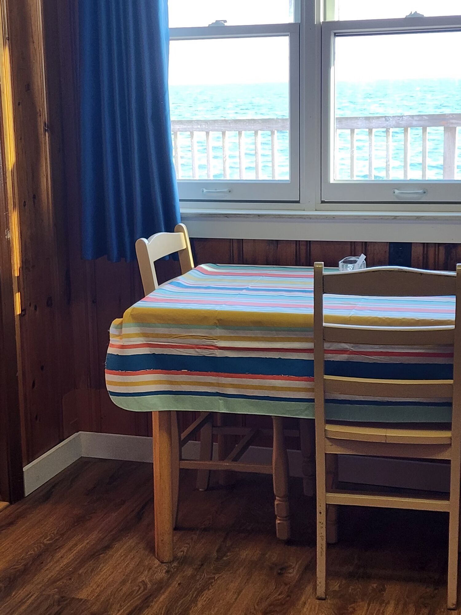 Una habitación con piso de madera, una mesa cubierta con mantel a rayas, una silla y vista al mar a través de una ventana con cortina.