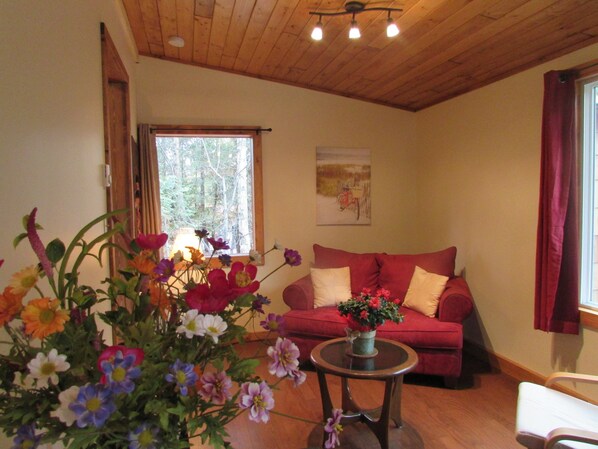 Un salon chaleureux avec un canapé rouge, une petite table ronde ornée d’un vase de fleurs et une fenêtre donnant sur les arbres.