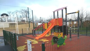 Een speelplaats met een rode glijbaan en klimconstructies, omgeven door een veiligheidshek.