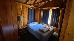 A wooden cabin bedroom with a bed, a nightstand, and a lamp. - (Cedar Cabin) Cozy 2 Bedroom Cottage - Waterfront - No Pets (East Tawas)