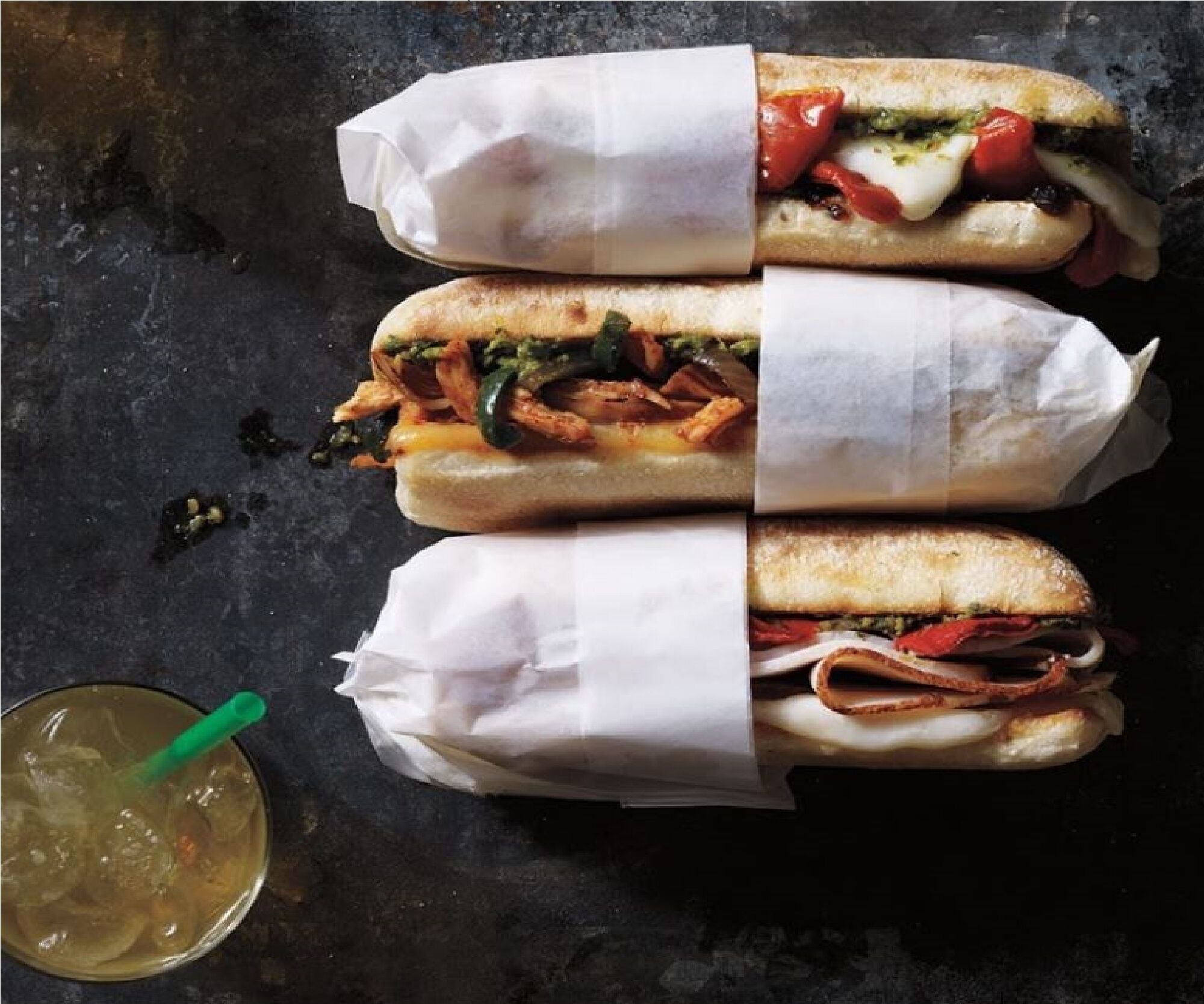 Three sandwiches wrapped in paper, a glass with a straw, and a dark surface.