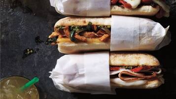 Three sandwiches wrapped in paper, a glass with a straw, and a dark surface.